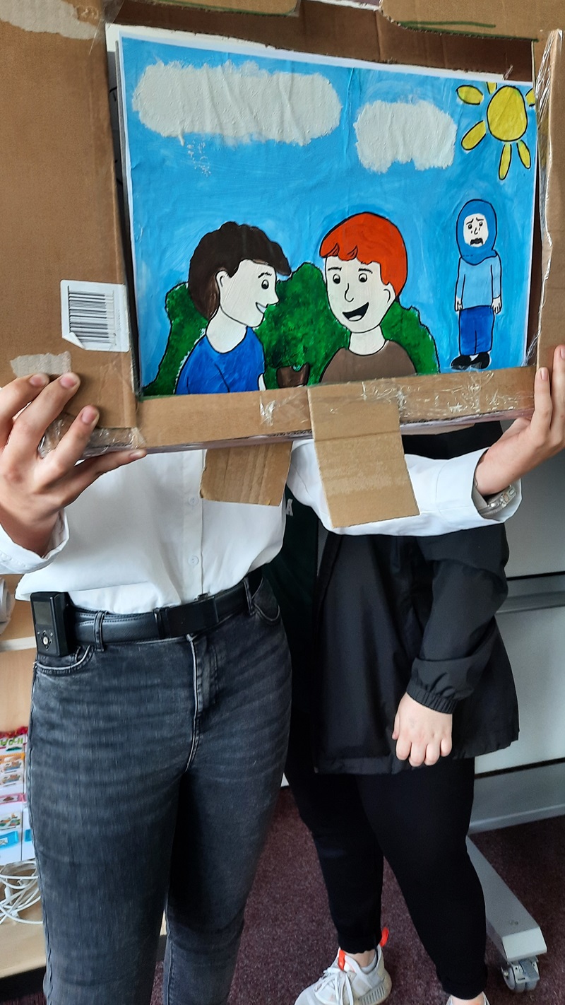 Sprache – ein Schlüssel zur Welt Selbst gestaltetes Kamishibai, das zwei gutgelaunte Menschen zeigt, die sich unterhalten, im Hintergrund eine traurige Frau mit Kopftuch, die aufgrund von Sprachbarrieren nicht richtig an der Gesellschaft teilhaben kann.