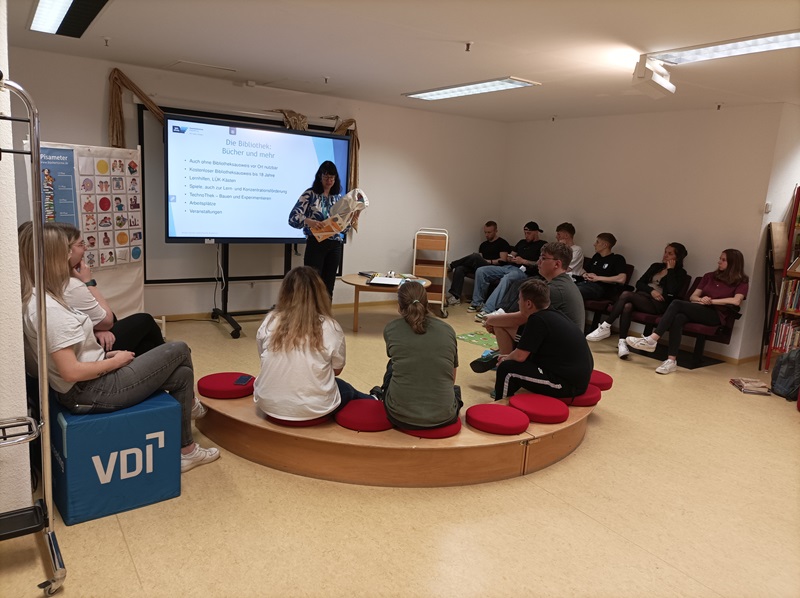 Sprache – ein Schlüssel zur Welt Die Auszubildenden der Euro Akademie Magdeburg sitzen in der Bibliothek auf Stühlen und Sitzen vor der Referentin und hören ihrem informativen Vortrag zu, der begleitet wird durch eine PowerPoint-Präsentation auf dem großen Whiteboard.