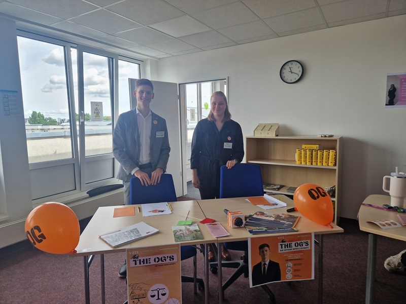Zwei Auszubildende stehen hinter einem Tisch in einem hellen Klassenraum mit großen Fenstern. Auf dem Tisch liegen orangefarbene Luftballons, Broschüren, Flyer, ein Klemmbrett, Stifte und weitere Unterlagen. Im Hintergrund steht ein Regal mit mehreren gelben Getränkedosen. An der Wand hängt eine Uhr, die kurz nach elf Uhr anzeigt. Die Personen tragen Namensschilder und formelle Kleidung.