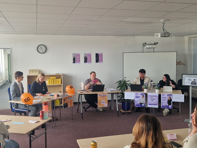 In einem hellen großen Klassenraum mit Teppichboden sitzen fünf Auszubildende der Euro Akademie Magdeburg an zwei Tischen, die im rechten Winkel zueinander stehen. Auf den Tischen stehen Laptops, Getränkedosen und orangefarbene Luftballons mit der Aufschrift 'OIC'. An den Tischen sind bunte Plakate mit Text und Logos befestigt. Im Hintergrund hängen drei lilafarbene Poster an der Wand, daneben steht ein Regal mit gelben Ordnern. Eine große weiße Tafel und eine Topfpflanze befinden sich ebenfalls im Raum. Weitere Schülerinnen und Schüler sitzen im Vordergrund und blicken zu den Tischen.