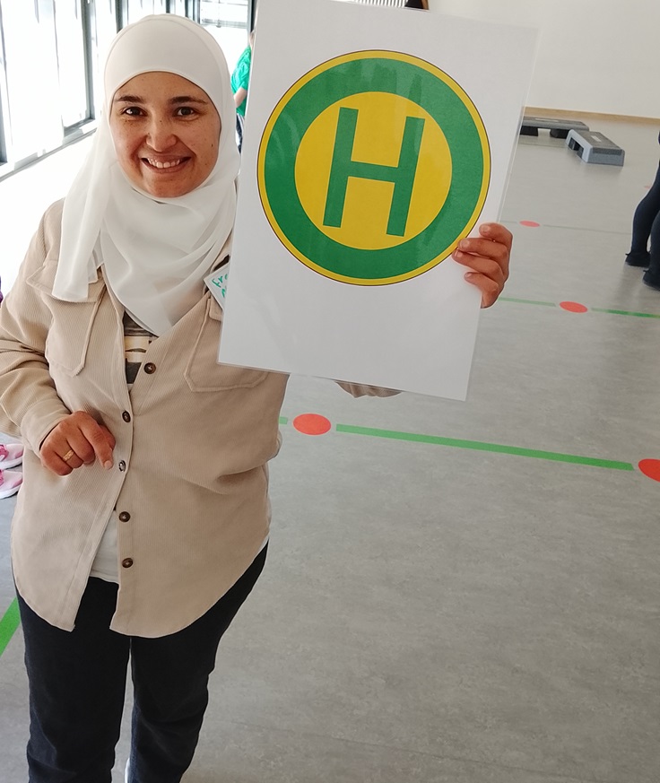 Eine Auszubildende mit beigefarbener Jacke und weißem Kopftuch hält ein Schild mit einem großen, grünen Kreis, in dessen Mitte ein gelbes Feld mit einem grünen Buchstaben 'H' zu sehen ist, das für Haltestelle steht. Der Hintergrund zeigt einen Raum mit grauem Boden, auf dem grüne Linien und rote Punkte markiert sind.