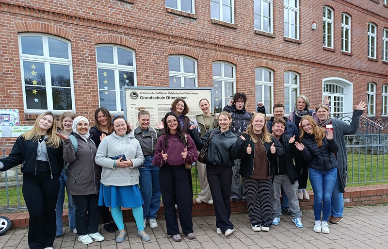 Vor einem Backsteingebäude mit großen Fenstern steht die Gruppe der Erzieherklasse der Euro Akademie Magdeburg. Die meisten Personen lächeln und zeigen mit erhobenen Daumen in die Kamera. Im Hintergrund ist ein Schild mit der Aufschrift 'Grundschule' zu erkennen. Einige Azubis tragen Jacken, andere Pullover oder Blusen. Die Gruppe steht auf einem gepflasterten Bereich vor einem Metallzaun. Das Wetter wirkt kühl, da viele langärmelige Kleidung tragen.