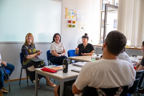 Sächsische Justizministerin zu Besuch an der Euro Akademie Meißen Der Blick aus dem Klassenraum in Richtung Tafel bzw. zeigt drei Frauen. Die rechte gestikuliert und scheint dabei zu sprechen. Der Blick der Frauen ist auf eine Person gerichtet, die nicht im Bild ist. Im Vordergrund rechts sieht man den Rücken eines Schülers in der ersten Reihe, der der Diskussion lauscht.