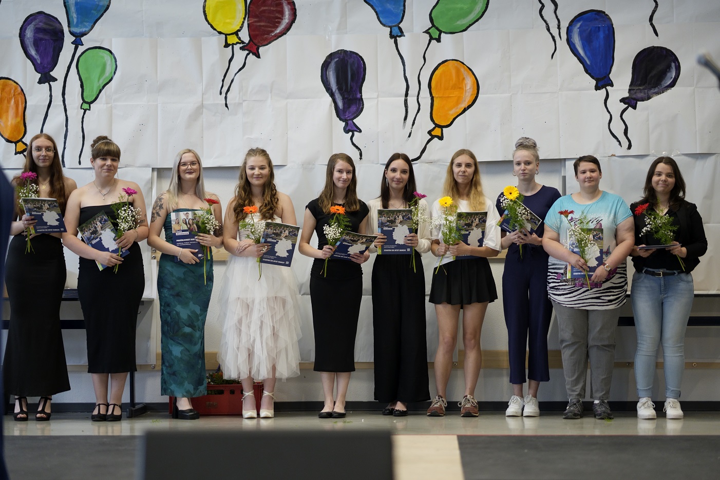 Zehn junge Frauen stehen in einer Reihe auf einer Bühne vor einem Plakat, auf dem bunte Luftballons gemalt sind. Sie tragen festliche Kleidung und in den Händen halten sie ein Dokument und Blumen.