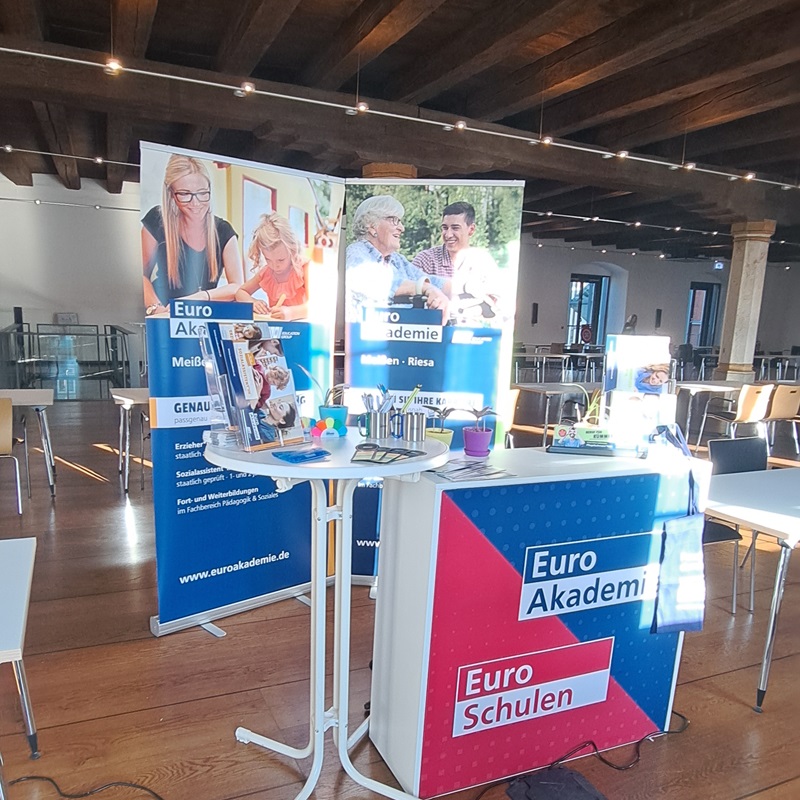 Messestand der Euro Akademie Meißen und Riesa in einem hellen Seminarraum mit Holzbalkendecke. Auf dem Stand sind Infobanner, Flyer und Werbematerialien zu Aus- und Weiterbildungen in Pädagogik und Sozialwesen zu sehen. Die Banner tragen die Aufschrift „Euro Akademie Meißen – Riesa“ und „Euro Schulen“. Der Raum ist modern eingerichtet mit Tischen und Stühlen für Besucherinnen und Besucher.