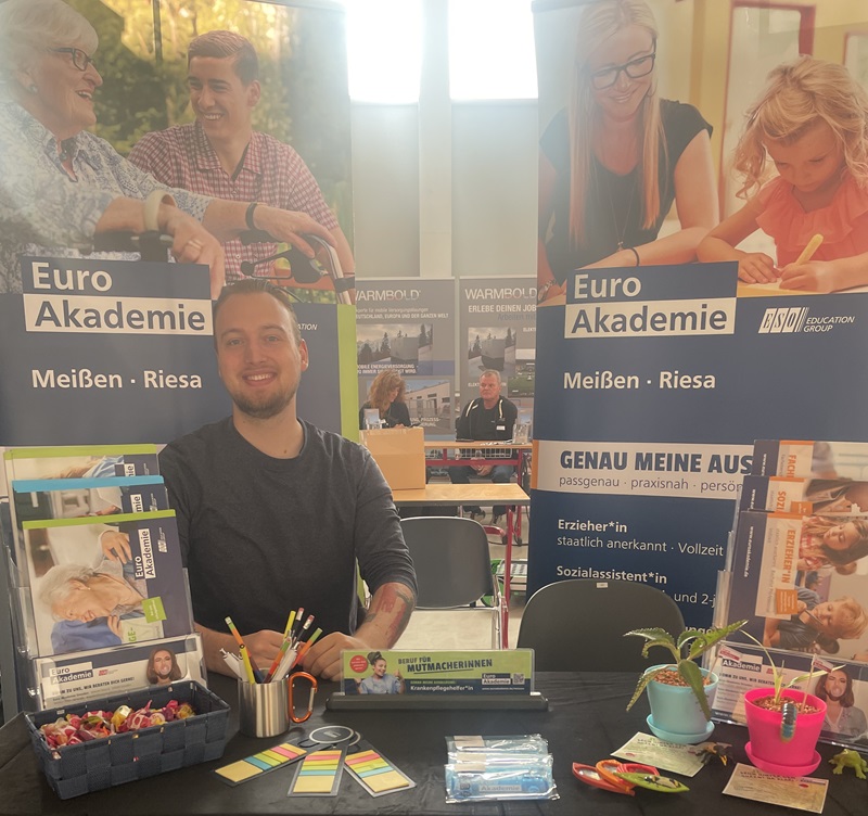 Ein junger männlicher Mitarbeiter sitzt gut gelaunt am Messestand der Euro Akademie und, der mit vielerlei Informationsmaterial ausgestattet ist, und lächelt freundlich für ein Bild in die Kamera. 
