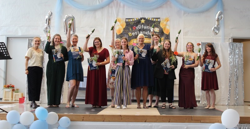 Eine Gruppe junger Absolventinnen steht auf einer Bühne und hält jeweils eine Rose und ein Dokument in der Hand. Sie tragen festliche Kleidung in unterschiedlichen Farben und Längen. Im Hintergrund hängt ein dekoriertes Banner mit Luftballons und der Aufschrift 'Prüfung Bestanden'. Die Bühne ist mit hellblauen und weißen Luftballons sowie silbernen Vorhängen geschmückt. Einige Frauen heben die Rose in die Höhe und lächeln.