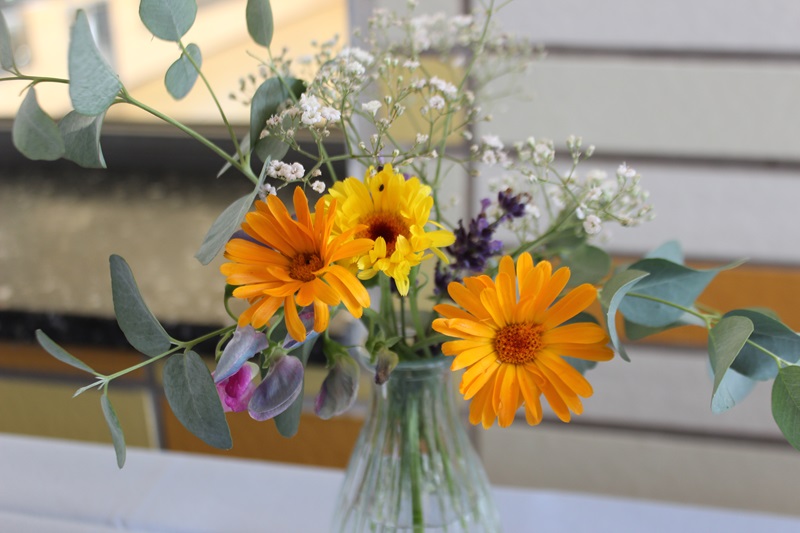 Ein Glasvase mit locker arrangierten Blumen steht auf einem weißen Untergrund. Im Strauß befinden sich orangefarbene und gelbe Blüten, einige kleine weiße Blüten sowie grüne, längliche Blätter. Im Hintergrund sind waagerechte, helle Paneele zu sehen.