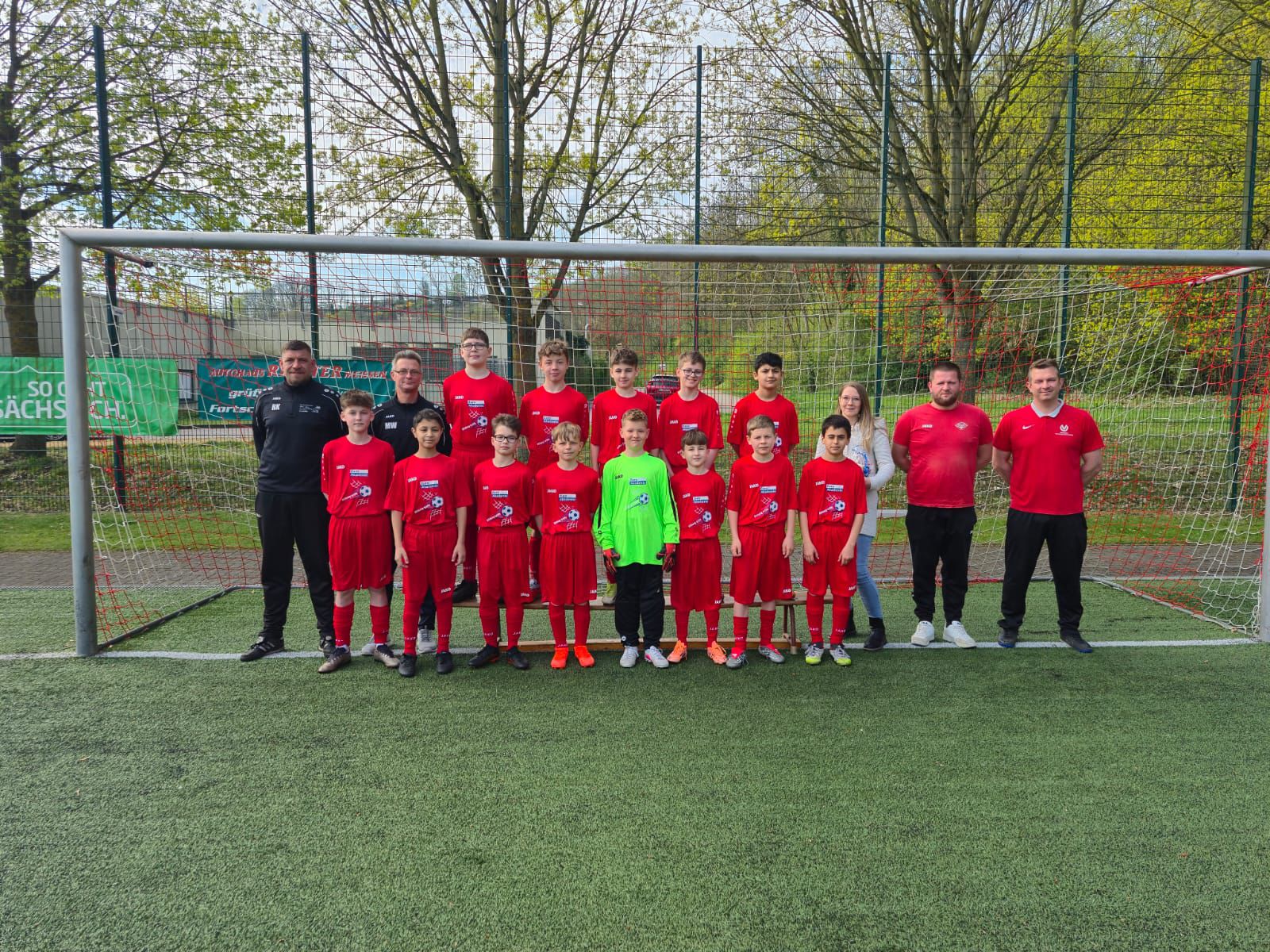 Vor dem Tor eines Fußballplatzes steht eine Jungenfußballmannschaft in roten Trikots zusammen mit ihren Trainern.