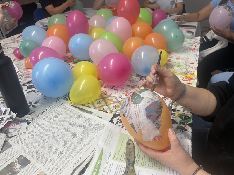 Im Vordergrund hält eine Person einen bunten Luftballon und beklebt ihn mit Papierstreifen, während sie einen Pinsel benutzt. Auf dem Tisch liegen zahlreiche weitere bunte Luftballons sowie Zeitungspapier und Bastelmaterialien. Im Hintergrund sitzen mehrere Personen und arbeiten ebenfalls mit Luftballons. Die Szene zeigt eine Bastelaktivität in einer Gruppe.