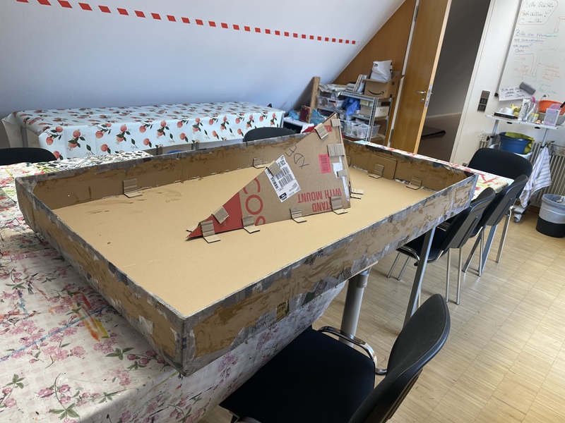 Ein großer rechteckiger Karton mit verstärktem Rand steht auf einem Tisch mit gemusterter Tischdecke. In der Mitte des Kartons steckt eine dreieckige Konstruktion aus Wellpappe mit aufgedrucktem Text und Barcode. Im Hintergrund befinden sich mehrere Stühle, ein Tisch mit einer weiteren Tischdecke und eine geöffnete Tür. An der Wand hängt ein Whiteboard mit handschriftlichen Notizen.