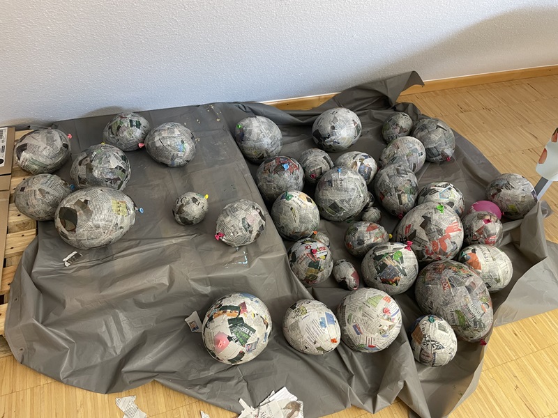 Auf einem hellen Holzboden liegt eine graue Plane, auf der zahlreiche papierüberzogene, eiförmige Objekte verteilt sind. Die Oberfläche der Objekte besteht aus Zeitungspapier, das teilweise farbige Ausschnitte zeigt. Einige der Eier sind mit kleinen bunten Papierstücken verziert. Im Hintergrund ist eine helle Wand zu sehen.