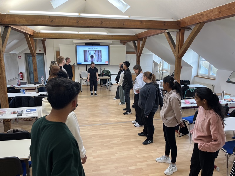 Auszubildende zur Pflegefachkraft der Euro Akademie Pößneck nehmen im Rahmen eines Gesundheitstages an einer praktischen Gruppenübung teil. Die Teilnehmenden hocken sich in zwei Reihen gegenüber auf den Boden und folgen den Anweisungen eines Trainers, der eine gesundheitsfördernde Übung demonstriert. Der lichtdurchflutete Seminarraum mit Holzstützen und Präsentationstechnik unterstreicht den praxisorientierten Charakter des Workshops. Die Szene vermittelt Teamarbeit, Bewegung, Gesundheitsprävention und Reflexion über den eigenen Umgang mit digitalen Medien.