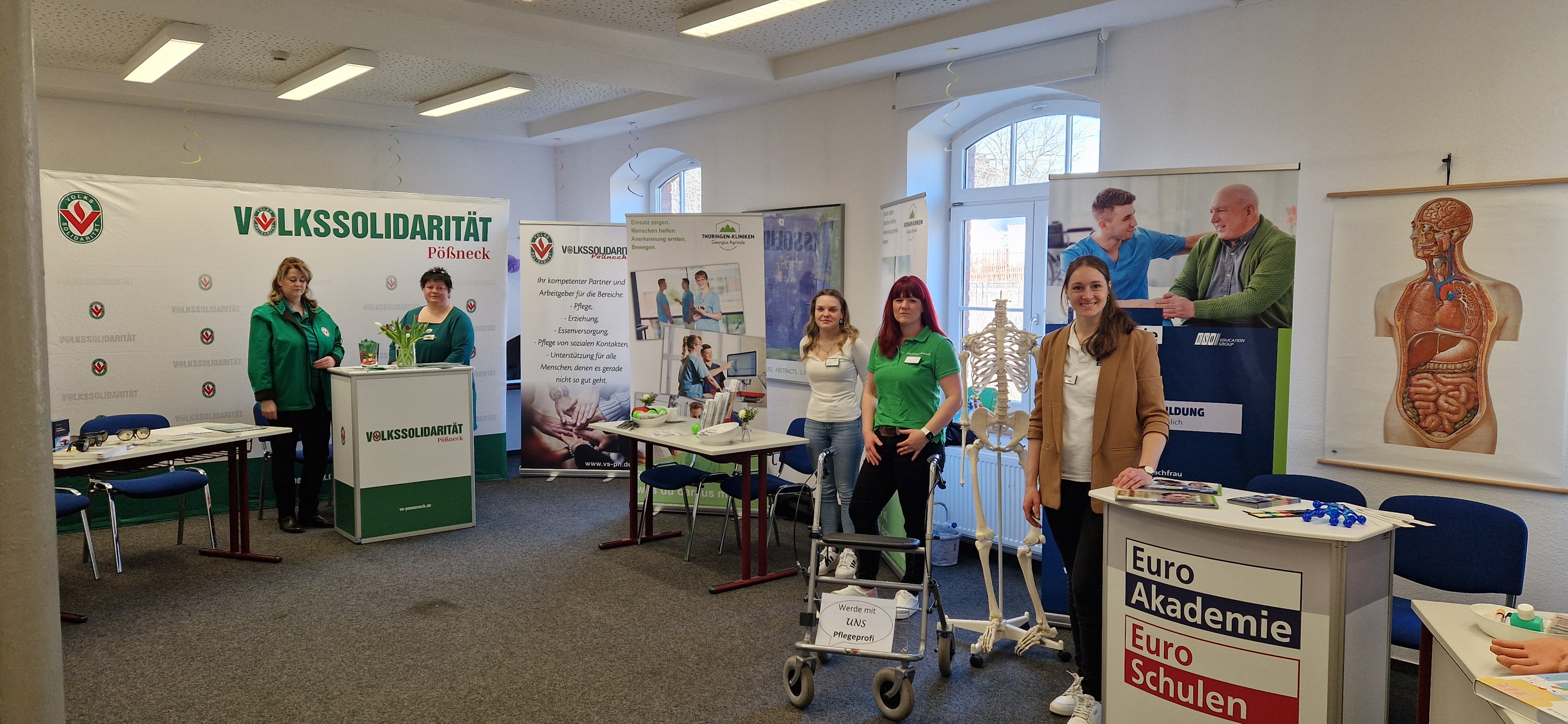 Ein Blick auf die gleiche Ausstellungsfläche aus einer leicht anderen Perspektive. Im Vordergrund ein Anatomie-Modell und ein Rollator mit dem Schild „Werde mit UNS Pflegeprofi“. Freundliche Standbetreuerinnen stehen bereit.