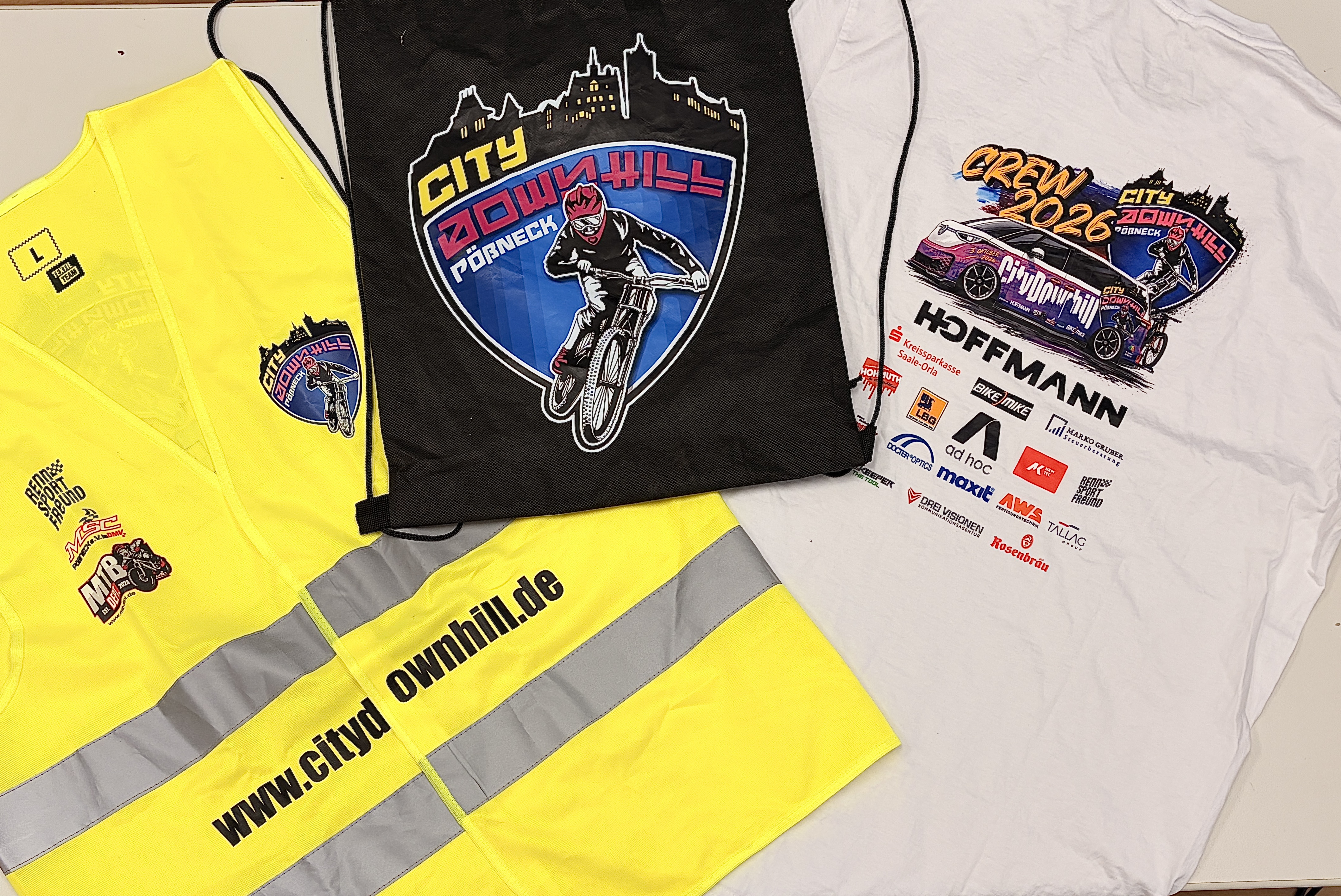 Gelbe Warnweste, schwarzer City-Downhill-Fößneck-Beutel und weißes Event-T-Shirt mit „Crew 2026“-Aufdruck und Sponsorenlogos liegen nebeneinander auf einem Tisch.