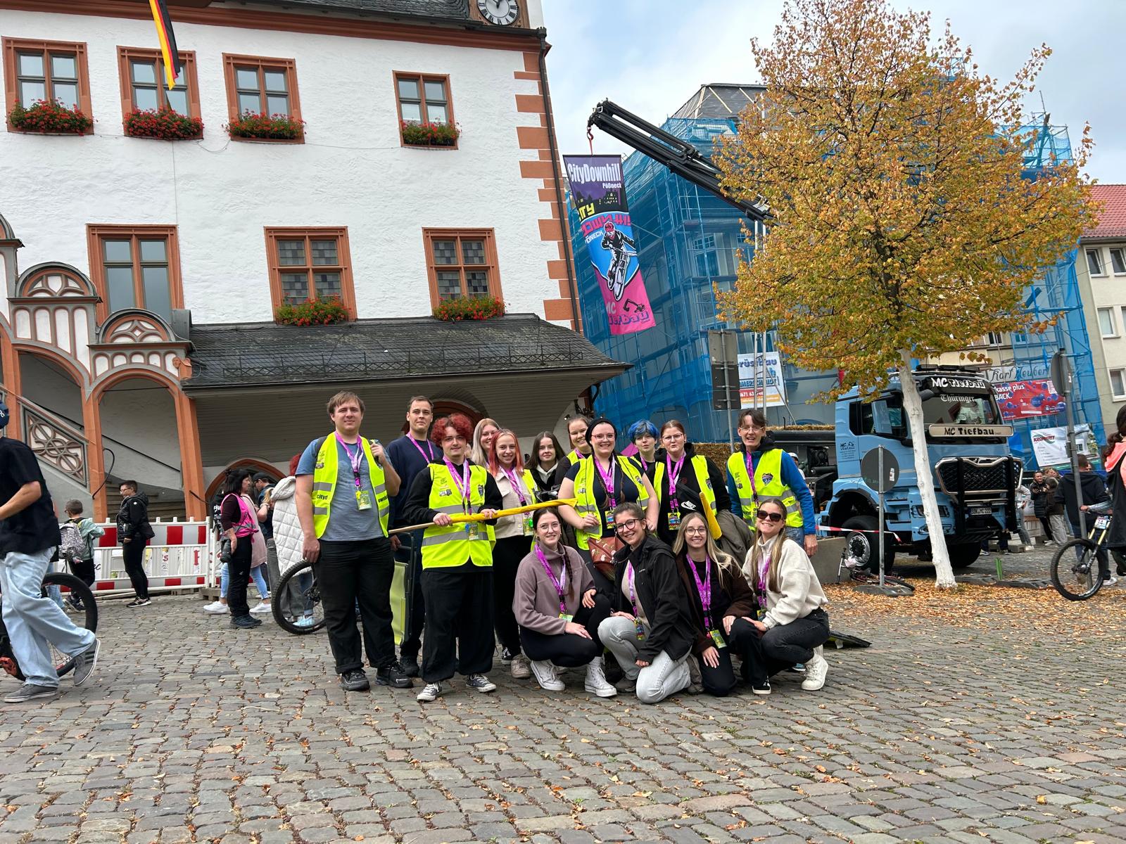 Gruppe von Helfer*innen und Teilnehmenden des City Downhill Fößneck posiert auf einem Marktplatz vor dem Rathaus. Mehrere Personen tragen gelbe Warnwesten und pinke Veranstaltungsbänder, im Hintergrund stehen Eventtechnik und ein Lkw.