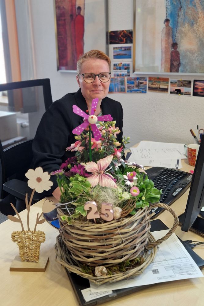Eine Person sitzt lächelnd an einem Schreibtisch in einem Büro. Vor ihr steht ein dekoratives Blumengesteck in einem Korb aus Weidenruten, das mit einer Windmühlen-Dekoration und einer Zahl ‚25‘ geschmückt ist. Im Hintergrund sind Bilder und Postkarten an der Wand zu sehen.