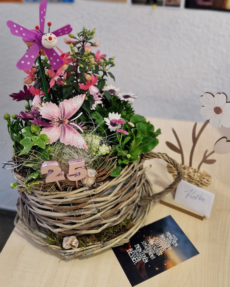 Ein dekoratives Blumengesteck in einem Korb aus Weidenruten, das mit einer Windmühlen-Dekoration und einer Zahl ‚25‘ geschmückt ist, steht auf einem Tisch. Daneben steht eine Deko-Blume aus Holz mit einem Schild, auf dem „Heike" geschrieben steht.