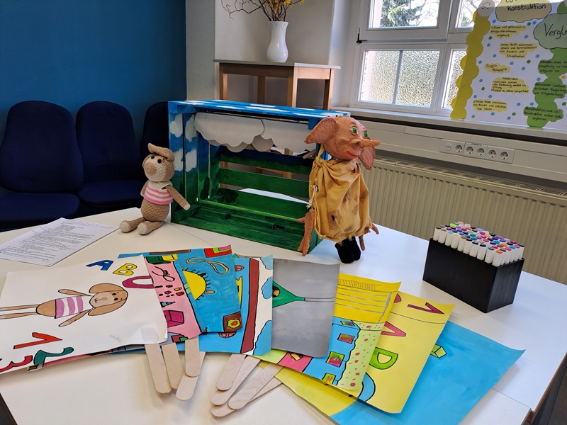 Auf einem Tisch liegen mehrere bunte Zeichnungen auf Papier, die mit Holzstäben versehen sind. Die Motive zeigen Buchstaben, Zahlen und kindliche Figuren. Daneben steht eine kleine grüne Kulisse aus Karton mit einer Bank, vor der eine Stoffpuppe mit rosa Kopf und gelbem Umhang platziert ist. Eine weitere Stoffpuppe sitzt links auf dem Tisch. Rechts befindet sich eine schwarze Box mit bunten Filzstiften. Im Hintergrund sind ein Fenster, eine Heizung und eine Pinnwand mit gelbem Papier sichtbar.