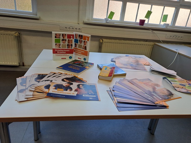 Auf einem weißen Tisch liegen mehrere Broschüren und Flyer, die farbig gestaltet sind. Die Broschüren zeigen Fotos von Menschen und enthalten Textfelder. Im Hintergrund steht ein aufgestellter Flyer mit mehreren Porträtfotos und farbigen Feldern. Die Tischfläche ist ordentlich, und im Raum sind weiße Fenster mit Milchglas sowie Heizkörper sichtbar.