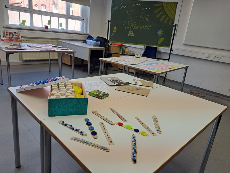 Ein Klassenzimmer mit mehreren Tischen, auf denen verschiedene Bastelmaterialien und Spiele verteilt sind. Auf einem Tisch liegen Holzstäbchen mit bunten Punkten, Flaschendeckel, Murmeln und kleine Behälter. Im Hintergrund steht eine grüne Tafel mit einer handschriftlichen Begrüßung und einer gezeichneten Sonne. Die Fenster lassen Tageslicht herein, und an den Wänden sind Heizkörper und Steckdosen sichtbar.