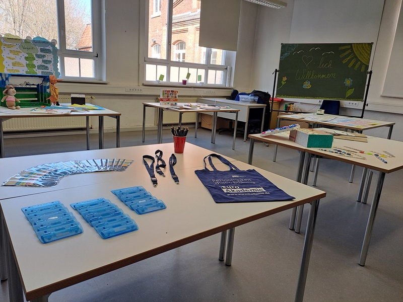 Mehrere Tische stehen in einem hellen Klassenraum. Auf den Tischen liegen verschiedene Gegenstände, darunter blaue Kunststoffboxen, Schlüsselbänder, ein blauer Stoffbeutel mit weißem Aufdruck, Fächer mit Informationsmaterial und ein roter Becher mit Stiften. Im Hintergrund sind große Fenster, eine grüne Tafel mit Kreidezeichnung und Schrift sowie weitere Materialien und Dekorationen zu sehen.