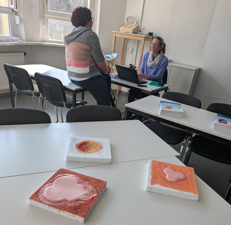 Zwei Personen sitzen an einem Tisch in einem hellen Raum mit mehreren weißen Tischen und Stühlen. Auf den Tischen liegen quadratische Platten mit farbigen, reliefartigen Formen, die an organische Strukturen erinnern. Im Hintergrund stehen Regale mit weiteren Gegenständen und ein Fenster lässt Tageslicht herein.