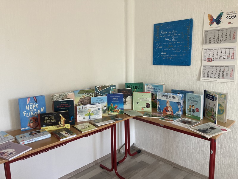 Blick auf zwei Tische, auf denen zahlreiche Literatur in Form von Büchern und Broschüren zum Thema Trauerarbeit mit Kindern ausliegt. An der Wand hängt ein blaues Bild mit einem Text, daneben ein Wandkalender.
