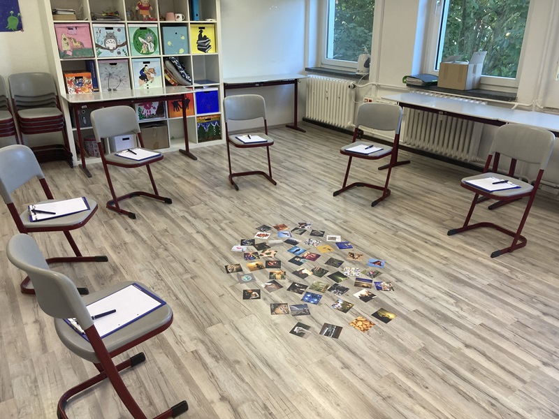 Sterben, Tod und Trauer – Kinder gut begleiten Blick in einen großen Raum, in dem ein Stuhlkreis aufgebaut ist, in der Mitte auf dem Boden liegen Bilder, im Hintergrund steht ein weißes Kallax-Regal, mit bunten Einschüben.
