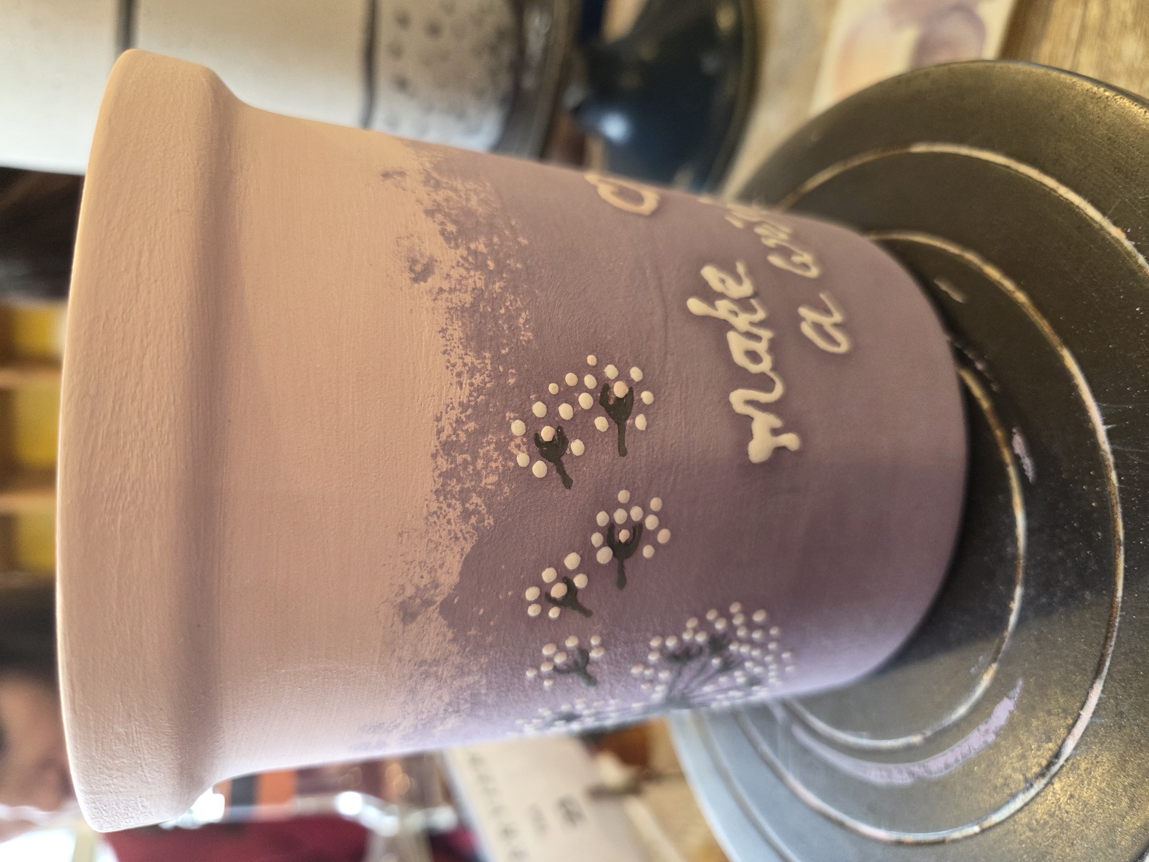 Nahaufnahme eines bemalten Keramikbechers in Lila-Verlauf mit weißen Punkten und Schriftzug; der Becher steht auf einer Drehscheibe im Keramikstudio.
