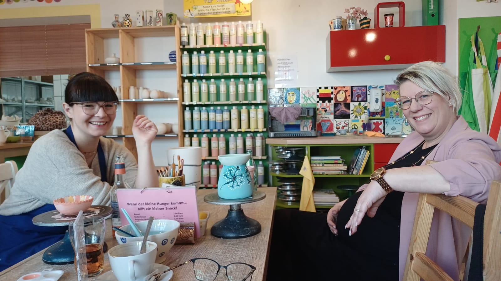 Zwei Frauen sitzen lächelnd an einem Tisch im Keramikstudio; vor ihnen stehen bemalte Keramikstücke auf Drehscheiben, Pinsel, Becher und Farbtöpfe.
