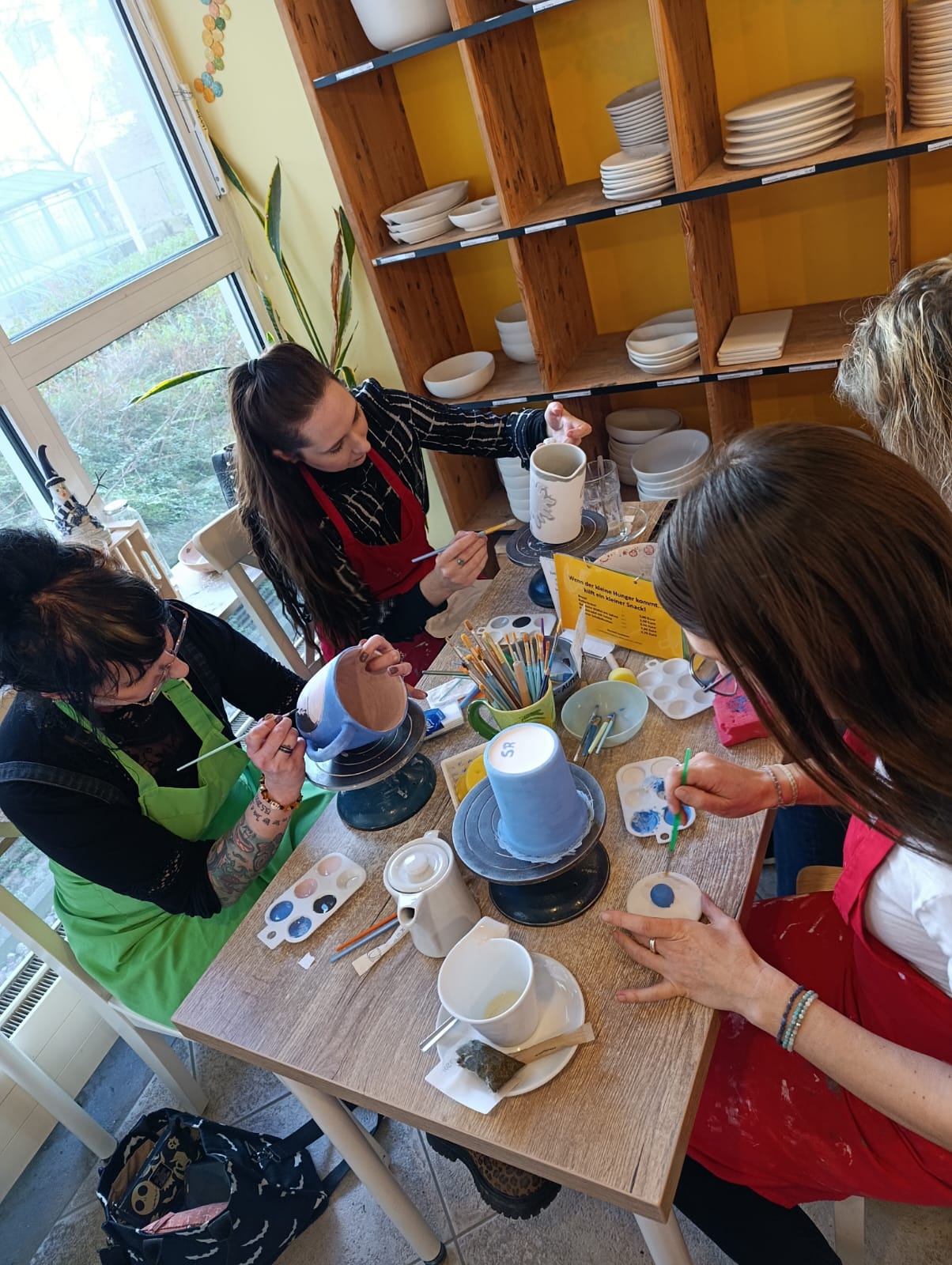 Mehrere Personen bemalen Keramik an einem Tisch; Paletten, Pinsel und Farbtöpfe liegen bereit, eine Person hält einen Keramikbecher über einer Drehscheibe.