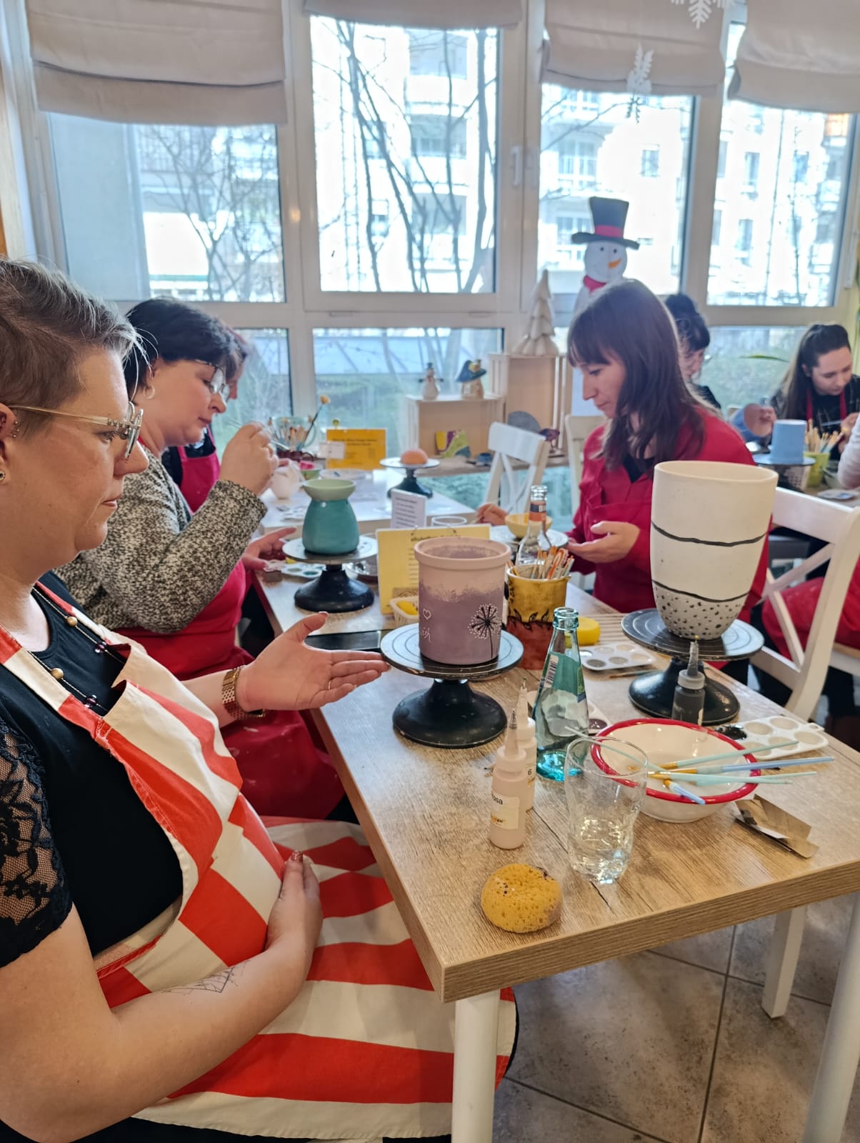 Mehrere Frauen bemalen Keramik in einem hellen Raum mit großen Fenstern; auf dem Tisch stehen Drehscheiben, Pinsel, Farben und Keramikbecher.