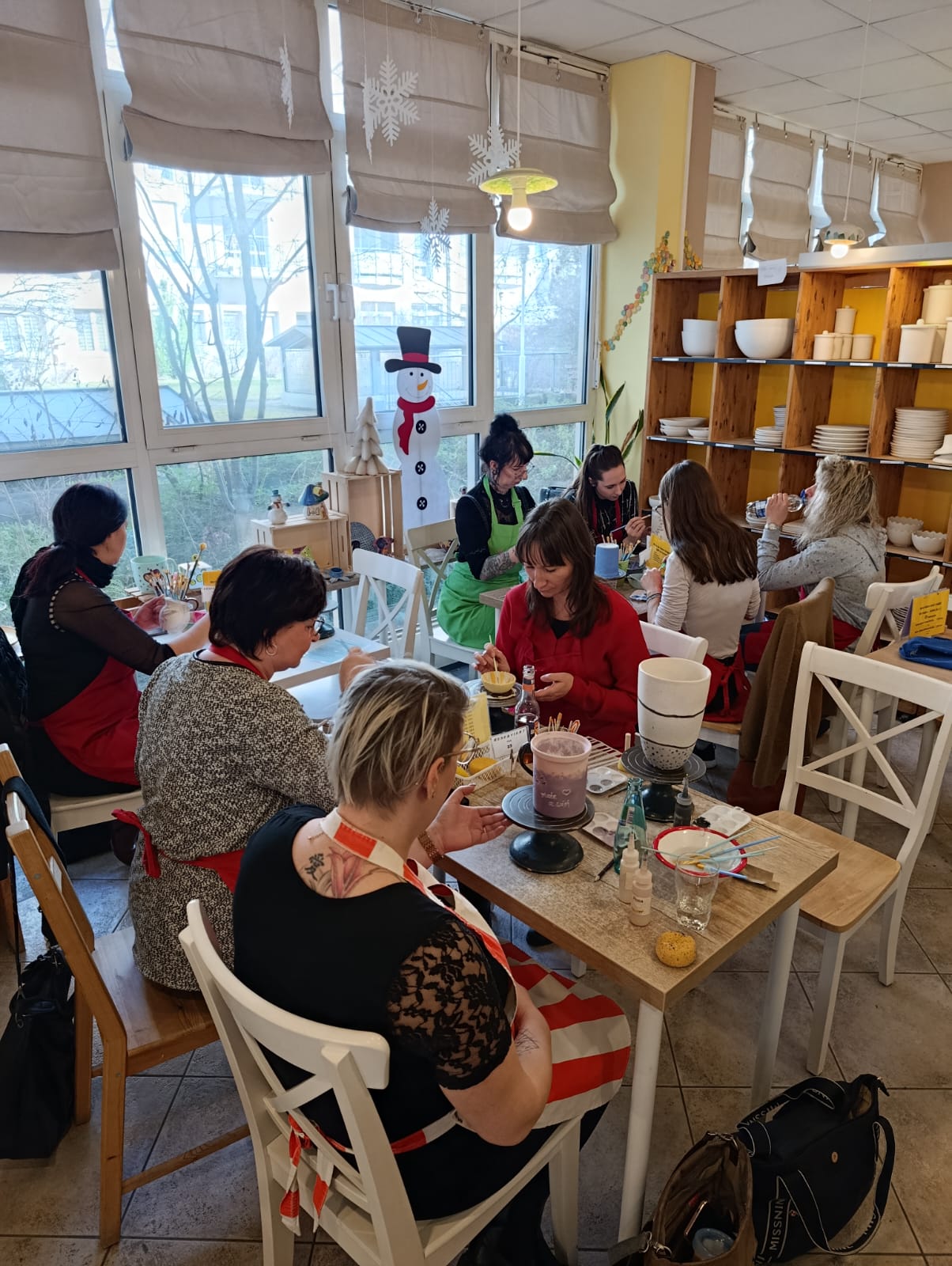 Große Gruppenansicht im Keramikstudio: mehrere Personen sitzen an Tischen und bemalen Keramik; im Hintergrund stehen Regale mit Schalen und Tellern sowie Winterdeko am Fenster.