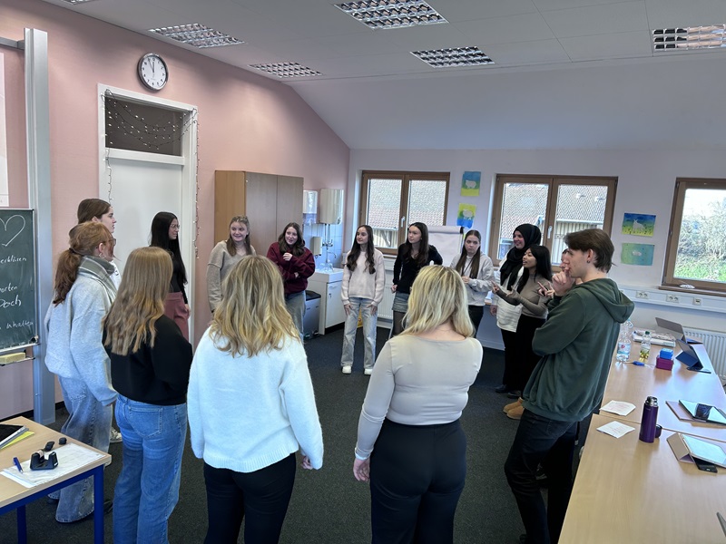 In einem hellen Klassenraum der Euro Akademie Tauberbischofsheim stehen mehrere Schüler*innen in einem lockeren Kreis zusammen. Die Personen tragen unterschiedliche, legere Kleidung, darunter Pullover und Jeans. Im Hintergrund sind große Fenster mit Blick auf eine Außenwand zu sehen, an der bunte Bilder hängen. An der linken Wand befindet sich eine Tafel mit Kreidezeichnungen und ein Uhr zeigt die Zeit. Der Raum ist mit Schränken und Tischen ausgestattet, auf denen verschiedene Gegenstände liegen. Die Szene vermittelt eine spontane Unterrichtssituation im Rahmen des „aktiven“ Adventskalenders, bei der die Schüler*innen eigenständig eine Unterrichtsstunde gestalten und durchführen.