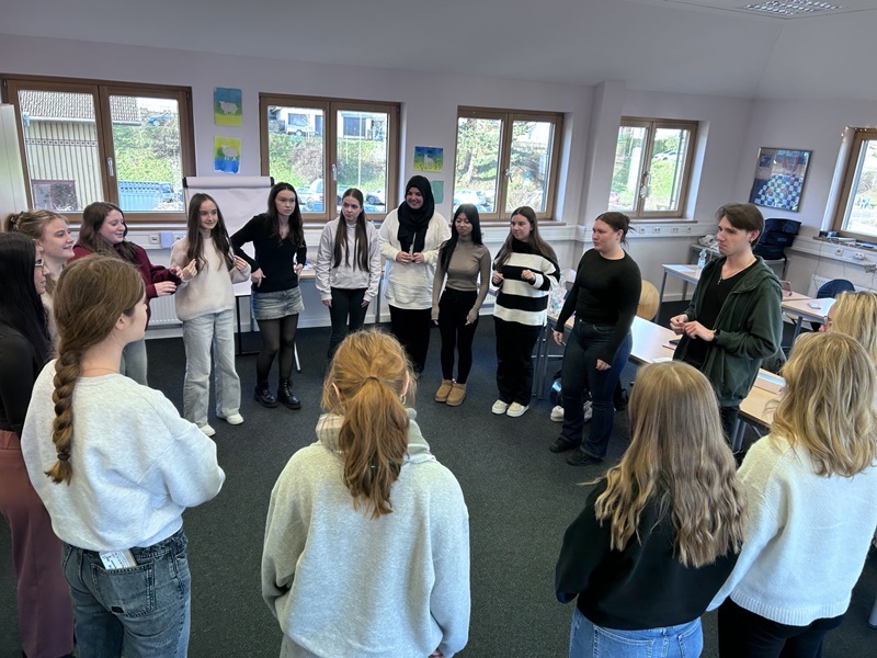 In einem hellen Klassenraum der Euro Akademie Tauberbischofsheim stehen mehrere Schüler*innen in einem lockeren Kreis zusammen. Die Gruppe besteht aus Jugendlichen, die legere Kleidung wie Pullover, Jeans und Strickjacken tragen. Die meisten haben lange Haare, einige sind zu Zöpfen geflochten. Die Wände sind mit bunten Bildern und Zeichnungen dekoriert, und durch die großen Fenster fällt Tageslicht herein. Die Szene zeigt eine spontane Unterrichtsstunde, die im Rahmen des „aktiven“ Adventskalenders organisiert wurde.