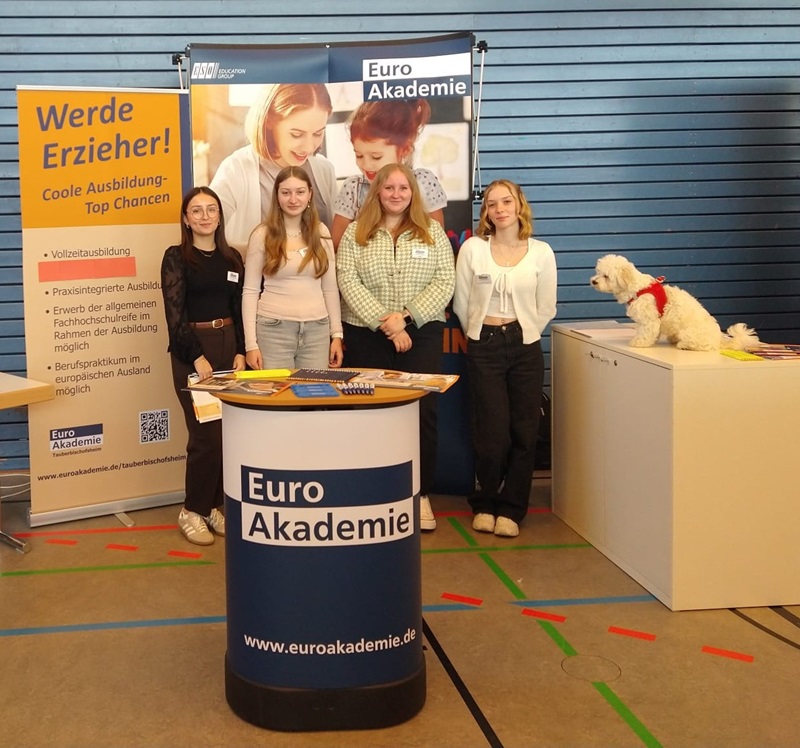 Im Vordergrund steht ein runder Messetisch mit dunkelblauer Verkleidung und dem Schriftzug 'Euro Akademie'. Dahinter stehen vier weibliche Auszubildende nebeneinander und posieren lächelnd für ein Gruppenbild. Sie tragen Freizeitkleidung und Namensschilder. Im Hintergrund hängt ein Roll-up-Banner mit dem Logo der Euro Akademie und weiteren Texten. Links davon steht ein gelbes Plakat mit Informationen zur Ausbildung als Erzieher. Rechts sitzt ein kleiner, weißer Hund mit rotem Halstuch auf einem weißen Podest. Der Boden ist mit buntem Klebeband markiert, die Wand besteht aus waagerechten blauen Paneelen.