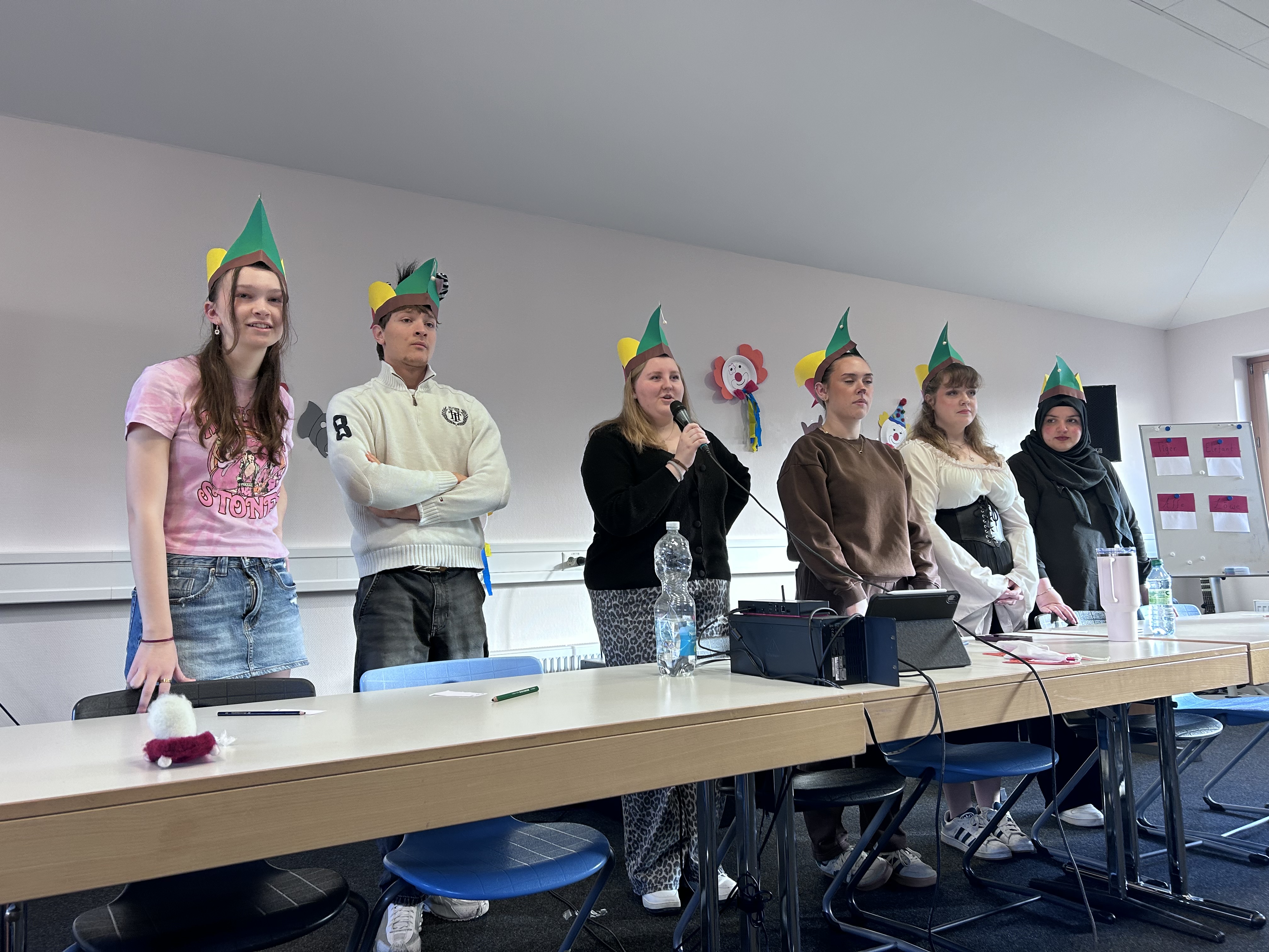 Sechs Schülerinnen und Schüler stehen hinter einem langen Tisch in einem Raum. Sie tragen zur Faschingsfeier grüne Papierhüte mit gelben und weißen Elementen. Auf dem Tisch liegen verschiedene Gegenstände, darunter eine Wasserflasche, ein Mikrofon, ein Laptop und Kabel. Im Hintergrund hängen bunte Papierdekorationen an der Wand. Die Personen stehen nebeneinander, einige mit verschränkten Armen, eine Person spricht in ein Mikrofon.