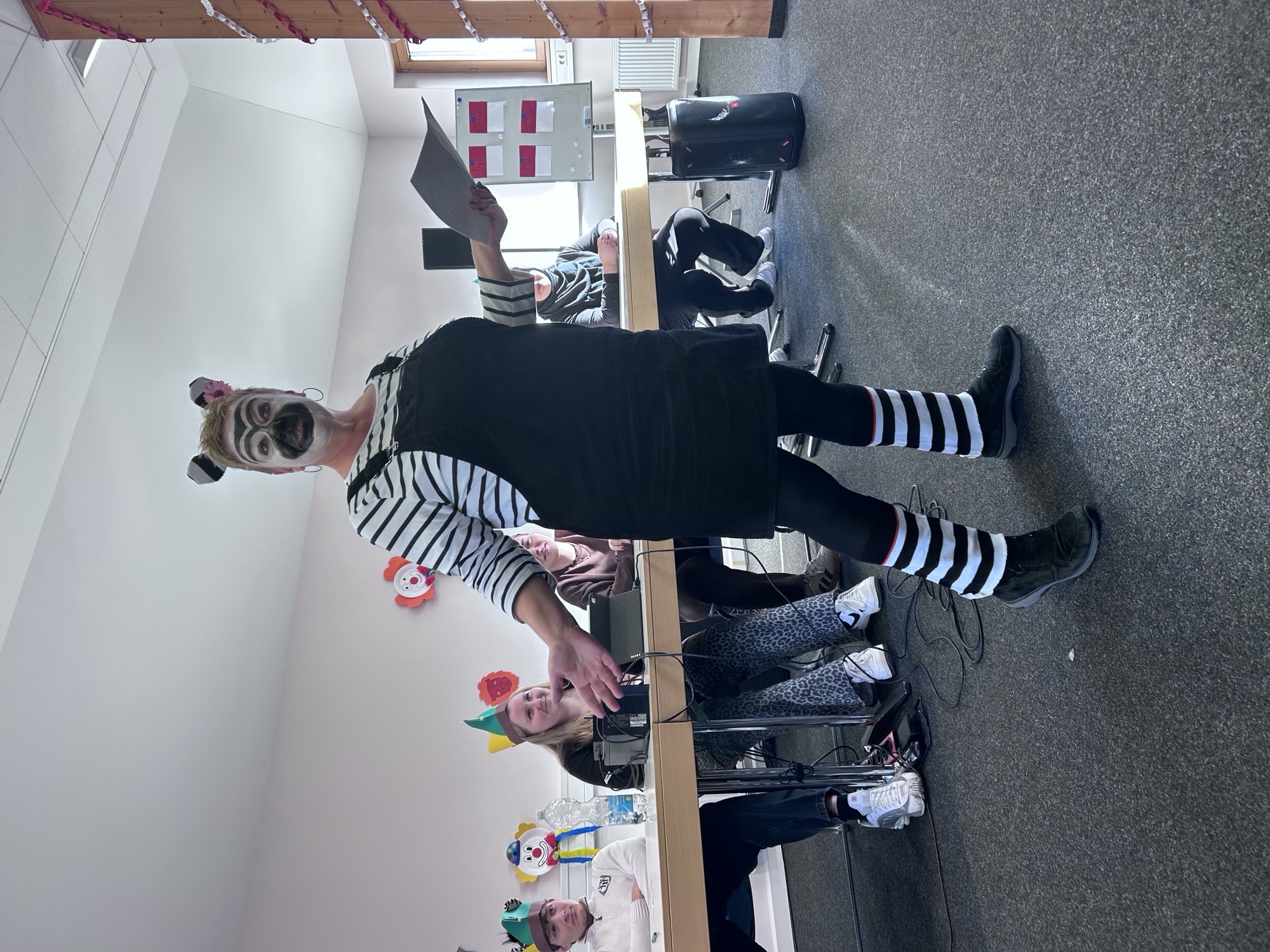 Eine Lehrerin steht in einem Raum und trägt ein gestreiftes Oberteil, einen schwarzen Latzrock, gestreifte Strümpfe und schwarze Schuhe. Sie ist als Zebra verkleidet. Am Kopf sind zwei kleine, runde Ohren befestigt. Im Hintergrund sitzen mehrere Personen an einem langen Tisch, einige tragen bunte Hüte oder Perücken. An der Wand hängen bunte Dekorationen. Die stehende Person hält ein Blatt Papier in der Hand und scheint zu sprechen oder etwas vorzutragen.