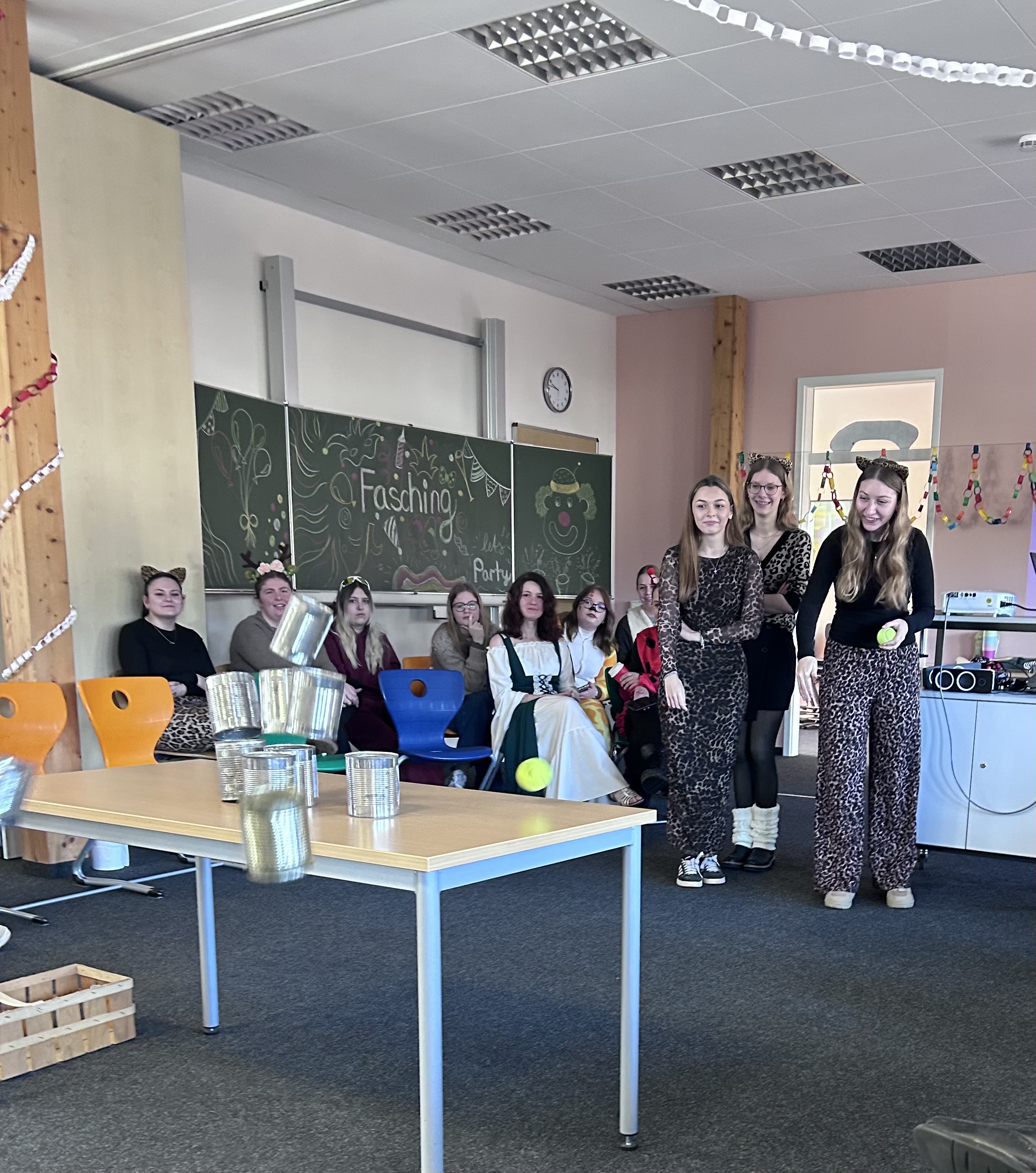 Eine Gruppe von Schülerinnen und Schülern befindet sich in einem festlich geschmückten Raum mit bunten Ketten und einer Tafel mit der Aufschrift „Faschingsfeier“. Drei Personen in Leopardenmuster-Kleidung stehen im Vordergrund, lächeln und halten einen Ball. Vor ihnen sind Metalldosen auf einem Tisch gestapelt, und ein Ball schwebt in der Luft und scheint sie umzuwerfen. Mehrere sitzende Personen, einige mit Kostümaccessoires wie Tierohren und Masken, beobachten das Geschehen.