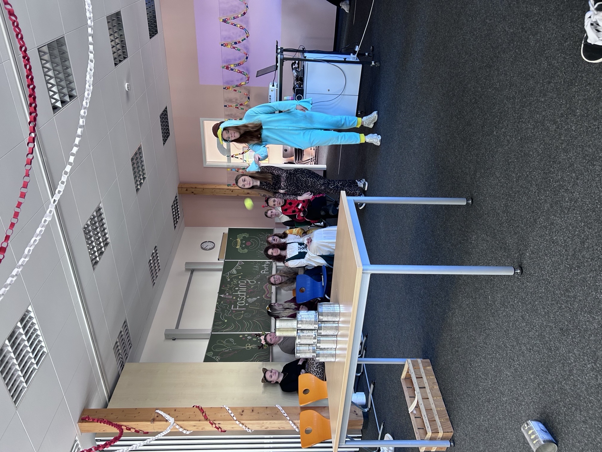 In einem festlich geschmückten Raum beobachtet eine Gruppe von Schülerinnen und Schülern ein Spiel, bei dem es darum geht, Dosen auf einem Tisch umzuwerfen. Zwei Personen stehen vorne, eine davon in einem blauen Overall, und wollen gerade einen Ball auf die Dosen werfen. Der Raum ist mit bunten Papiergirlanden dekoriert, und im Hintergrund ist eine Tafel mit der Aufschrift „Fasching“ zu sehen. Die Szenerie vermittelt eine festliche oder partymäßige Atmosphäre.