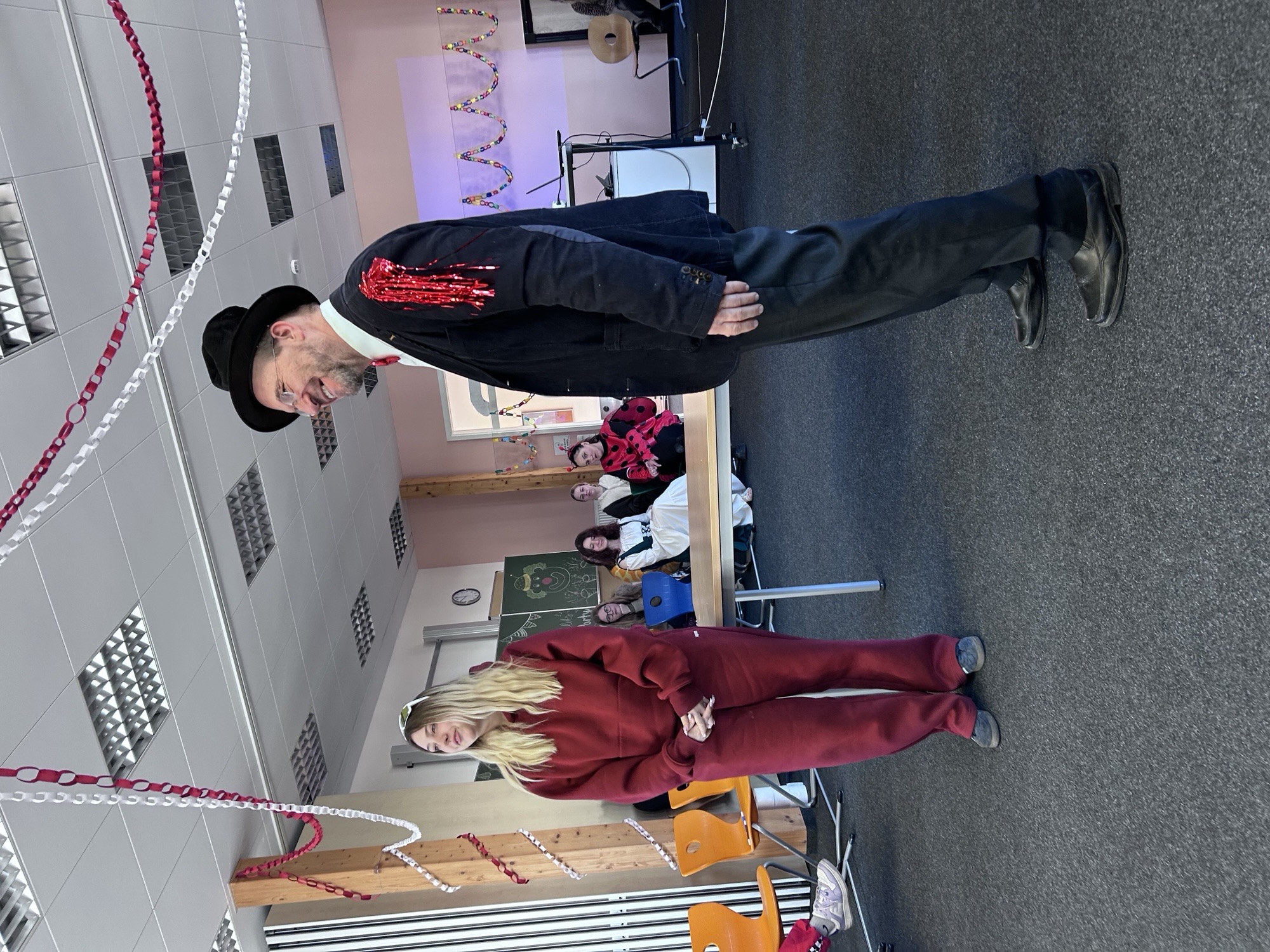 Ein Lehrer und eine Schülerin stehen in einem festlich geschmückten Raum zur Faschingsfeier. Die Schülerin trägt einen roten Trainingsanzug, der Mann ein Sakko mit glitzernden roten Quasten an den Schultern, einen Zylinder und eine Krawatte. Der Raum ist mit rot-weißen Papiergirlanden dekoriert, und im Hintergrund sitzen mehrere Personen, einige davon kostümiert. Die Atmosphäre wirkt festlich.