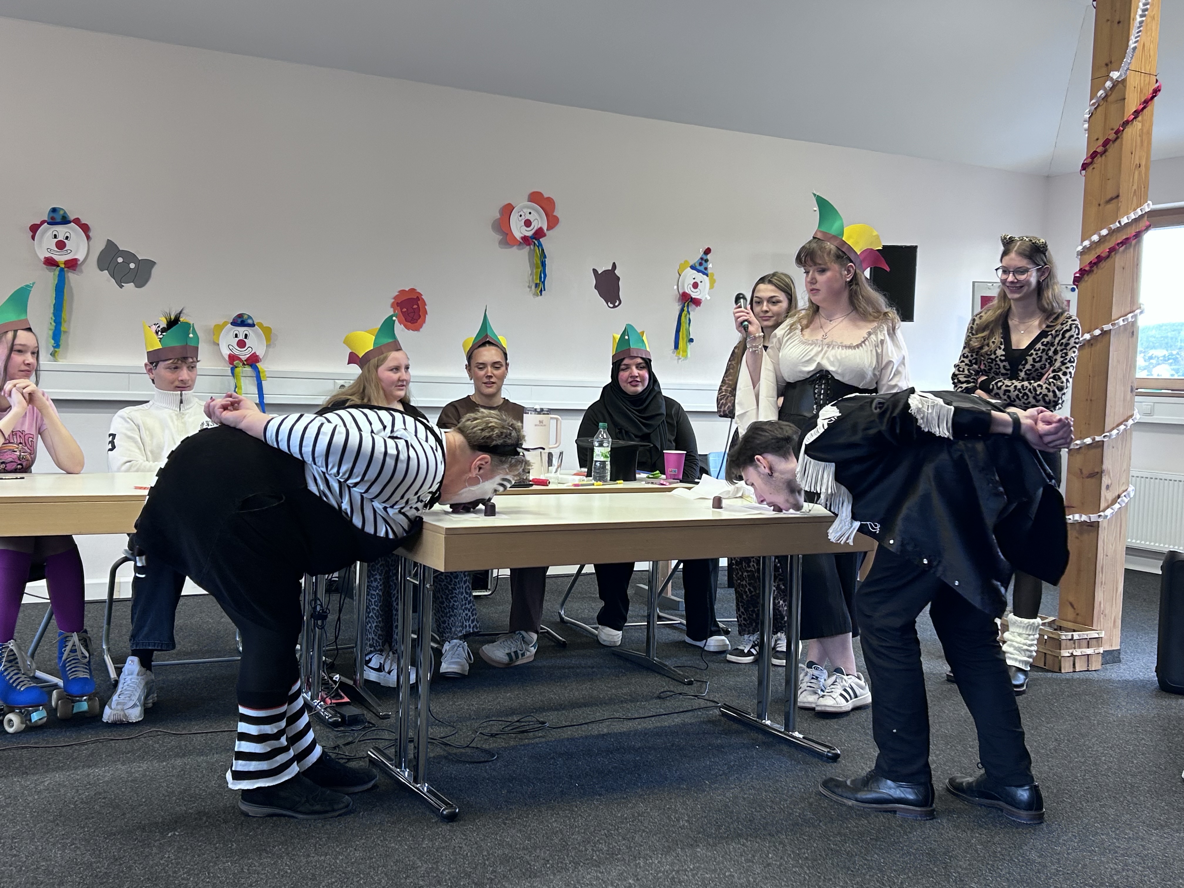 Das Bild zeigt einen festlich geschmückten Raum, in dem sich die verkleideten Schülerinnen, Schüler und Lehrkräfte vergnügen. Zwei Personen im Vordergrund, verkleidet, beugen sich mit auf dem Rücken gehaltenen Händen über einen Tisch und spielen Wettessen von Schokoküssen. Eine weitere Gruppe sitzt an den Tischen und trägt bunte Hüte, die an einen Zirkus oder eine Party erinnern. Die Wände sind mit Clown- und Tierfiguren dekoriert, die die lebhafte und fröhliche Atmosphäre unterstreichen.