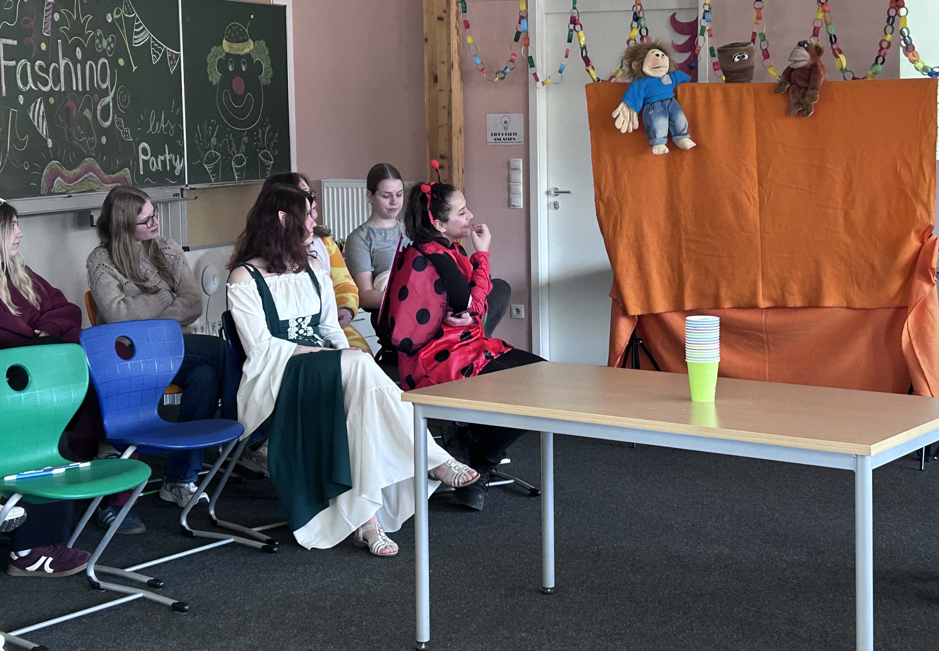 Die Schülerinnen sitzen in einem für die Faschingsfeier geschmückten Klassenraum. Einige sind verkleidet, darunter ein Marienkäfer und jemand in einem mittelalterlichen Kleid. Sie beobachten ein Puppentheater hinter einem orangefarbenen Tuch. Zur Dekoration gehören bunte Papiergirlanden und eine Tafel mit Zeichnungen und den Worten „Fasching“ und „Lasst uns feiern“. Im Vordergrund steht ein Tisch mit gestapelten grünen Bechern.