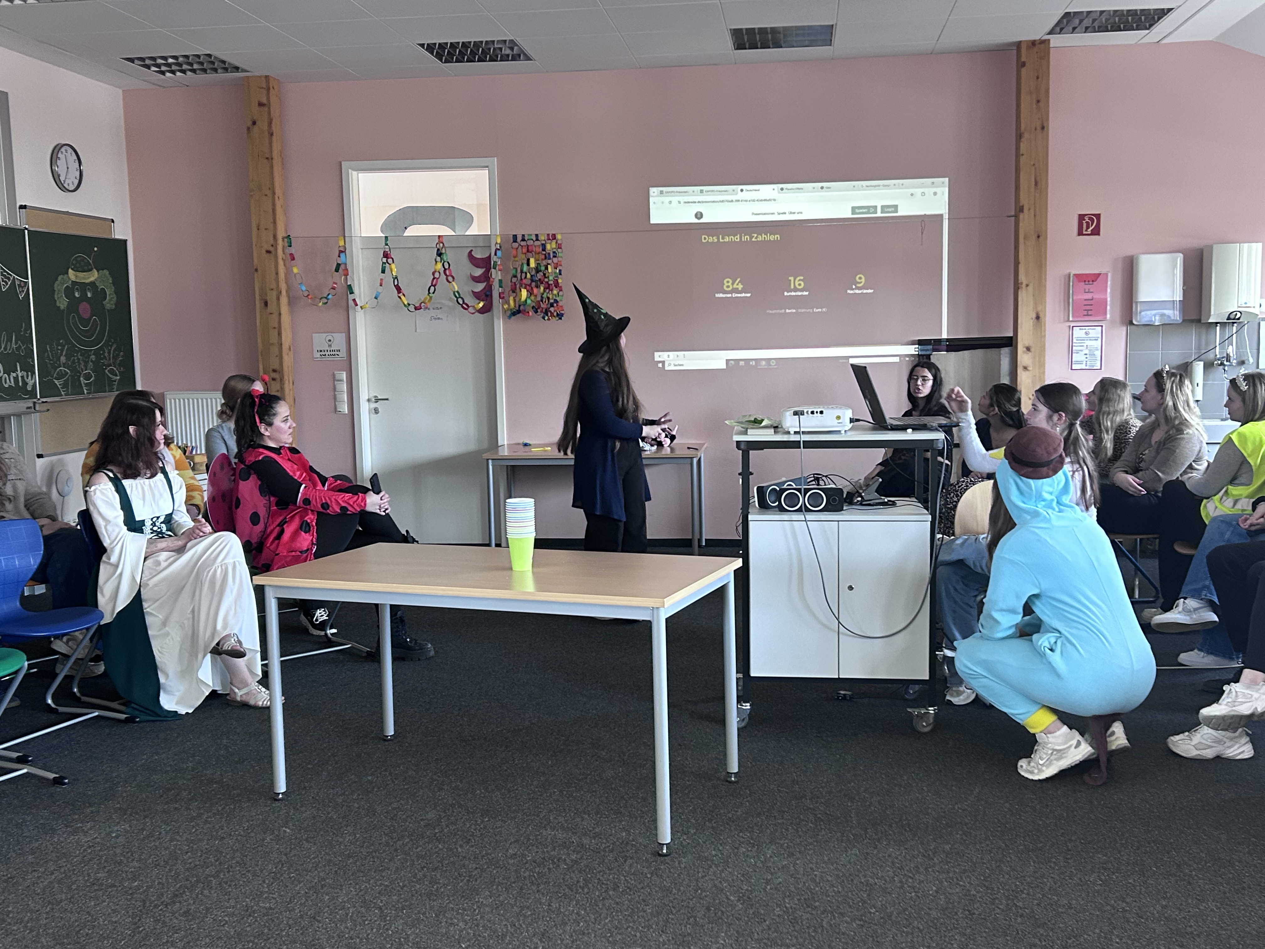 Eine Klassenzimmerszene mit verkleideten Schülerinnen und Schülern, die sitzen und stehen, während jemand mit einem Beamer präsentiert. Der Raum ist mit einer bunten Papiergirlande und Zeichnungen an der Tafel geschmückt. Zu den Kostümen gehören ein Marienkäfer, eine Hexe, ein mittelalterliches Kleid und ein blauer Tieranzug. Die Atmosphäre ist festlich im Sinne der Faschingsparty.