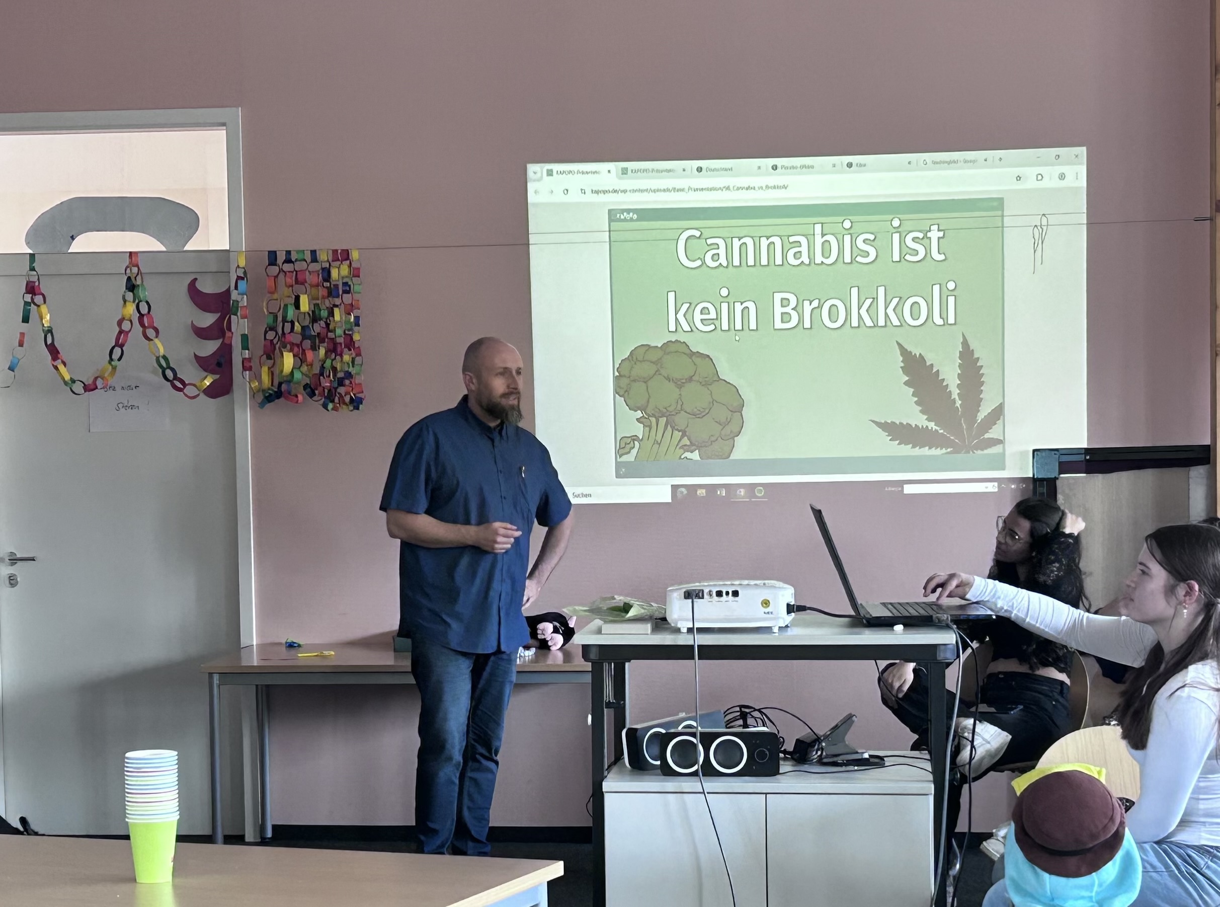Ein Lehrer hält in einem Klassenzimmer einen Vortrag. Der Beamer zeigt eine Folie mit dem deutschen Text „Cannabis ist kein Brokkoli“, begleitet von Abbildungen von Brokkoli und einem Cannabisblatt. Zwei Schüler sitzen an einem Tisch mit Laptop und Lautsprechern. Der Raum ist, im Rahmen der Faschingsfeier, mit bunten Papiergirlanden geschmückt. Auf einem nahegelegenen Tisch stehen Plastikbecher.