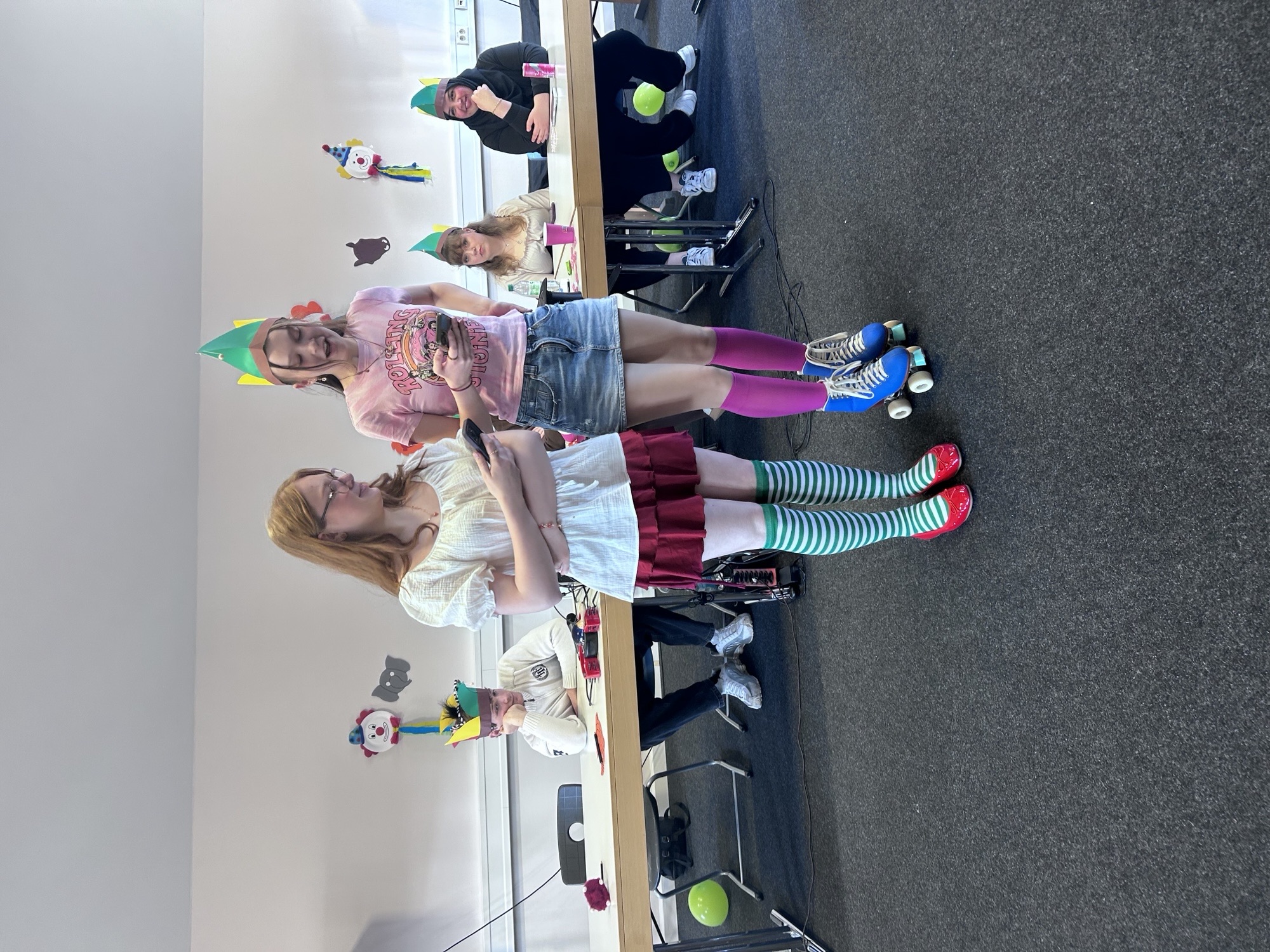 Zwei Schülerinnen stehen vor einem mit Luftballons und Papierbastelarbeiten geschmückten Tisch. Sie tragen bunte Hüte und unterschiedliche Socken, eine von ihnen fährt Rollschuhe. Hinter ihnen sitzen weitere Schülerinnen und Schüler, ebenfalls mit Hüten. Der Raum ist zur Faschingsfeier festlich geschmückt.