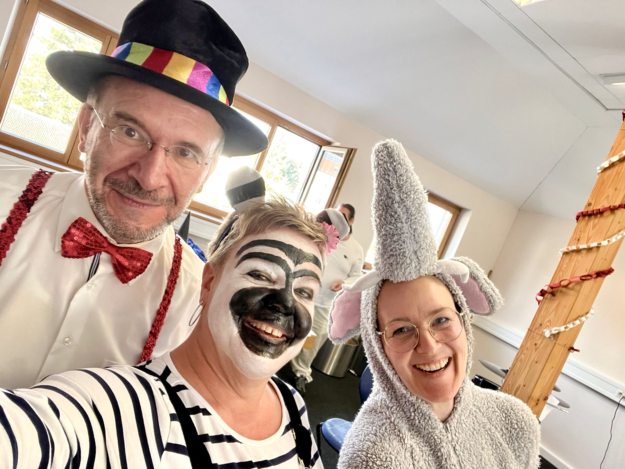 Drei Personen tragen bunte Verkleidungen in einem hellen Raum. Links ist ein Mensch mit schwarzem Hut mit Regenbogenband, roter Fliege und roten Hosenträgern zu sehen. In der Mitte steht eine Person mit schwarz-weiß gestreiftem Oberteil. Rechts trägt jemand ein graues Elefantenkostüm mit großem Rüssel und Ohren. Im Hintergrund sind Fenster und ein Holzpfosten mit rot-weißer Kordel sichtbar.