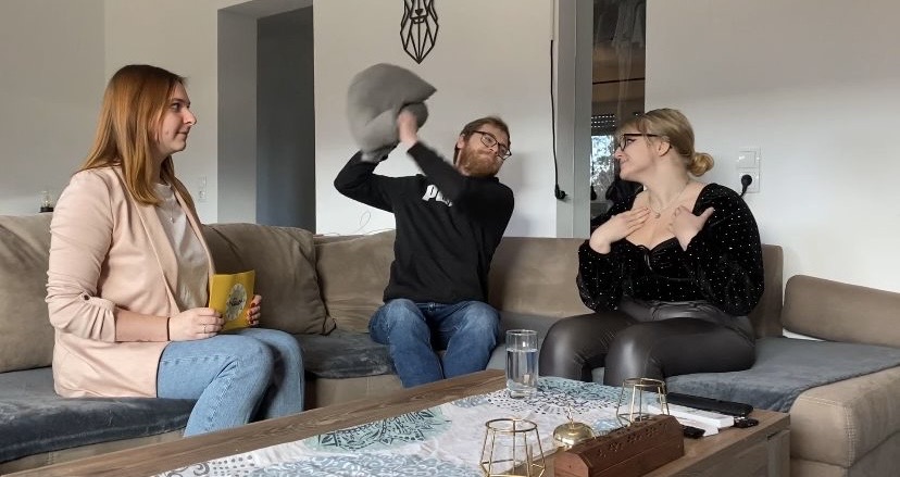In diesem Bild sind drei Personen in einem Wohnzimmer zu sehen. Eine Frau mit langen, roten Haaren sitzt auf einem Sofa und hält ein Karte in der Hand. Ein Mann mit Brille und Bart sitzt in der Mitte des Sofas und hält ein graues Kissen über seinem Kopf, als würde er es gerade werfen oder halten. Eine weitere Frau mit blonden Haaren, die zu einem Dutt gebunden sind, sitzt auf dem Sofa und hat ihre Hände auf der Brust verschränkt. Vor ihnen steht ein Couchtisch mit einigen Gegenständen darauf, darunter Gläser und eine Flasche. Der Raum wirkt gemütlich und ist modern eingerichtet.