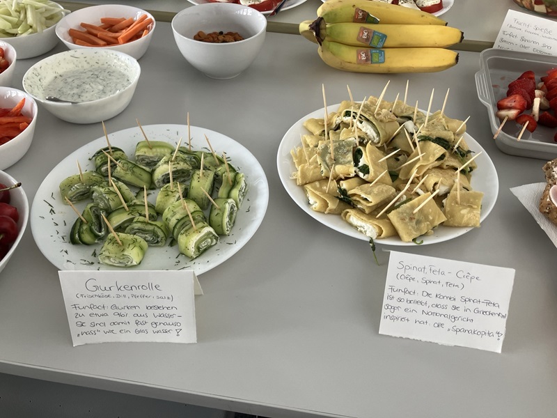 Auf Tellern angerichtet sind Gurkenrollen, Spinat-Feta-Crêpes, Bananen, in Schälchen ein Kräuterdip, daneben in Streifen geschnittene Paprika und Karotten.